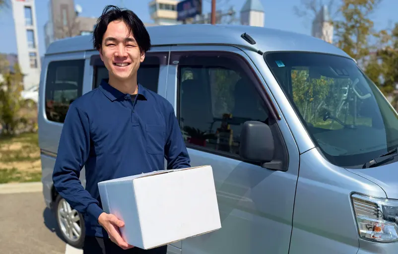 軽バンの前で荷物を持ち笑顔の軽貨物ドライバー